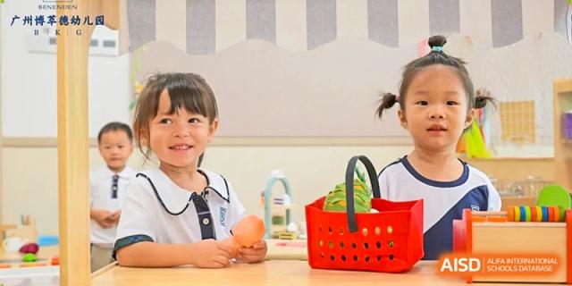 Two smiling young students stand behind a play kitchen set with a red basket during a creative role-play session at Benenden Bilingual School Guangzhou