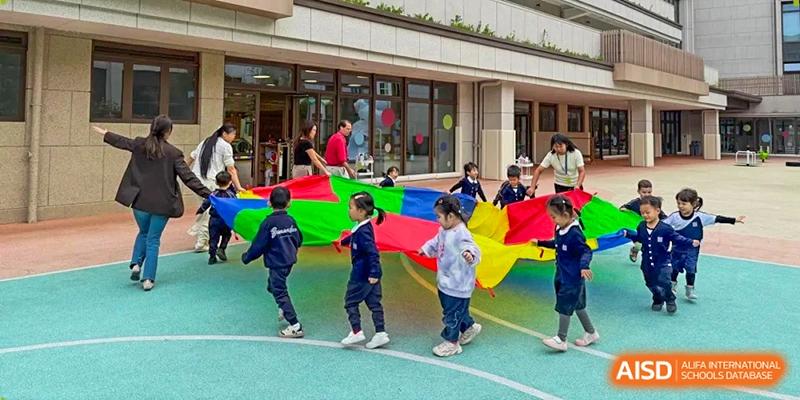 Young children play with a large, colorful parachute during an outdoor physical education activity on campus at Benenden Bilingual School Guangzhou