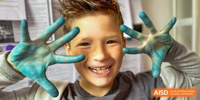 A smiling young boy proudly shows off his hands covered in bright blue paint during an art activity. Deutsche Botschaftsschule Peking