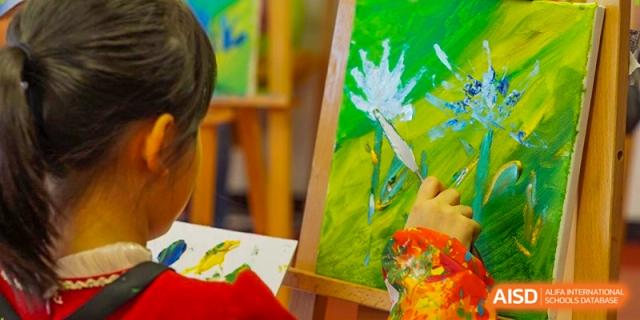 A student uses a palette knife to apply vibrant green and yellow paint to a canvas on an easel during art class. Deutsche Botschaftsschule Peking