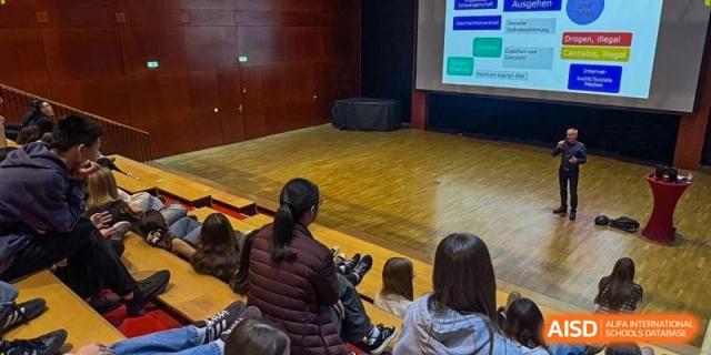 Students sit in an auditorium facing a large projector screen displaying an educational presentation. Deutsche Botschaftsschule Peking