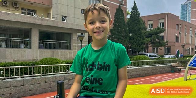 A young boy wearing a green t-shirt smiles brightly while sitting outside on a sunny day at school. Deutsche Botschaftsschule Peking