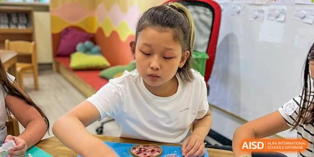 A young student works diligently on a colorful craft project at her desk in a bright classroom setting. Deutsche Botschaftsschule Peking