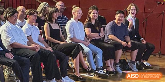 A row of students and teachers sit in an audience, smiling and engaged during a school performance. Deutsche Botschaftsschule Peking