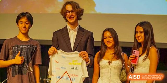 Two students pose proudly on stage while holding a signed t-shirt as a memento of their time at school. Deutsche Botschaftsschule Peking
