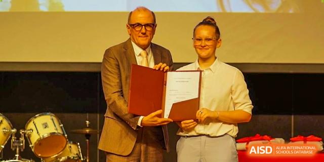 A smiling graduate in a white shirt receives her official certificate during the formal graduation event. Deutsche Botschaftsschule Peking