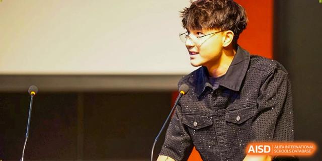 A student wearing glasses speaks into a microphone while addressing the audience during a school event. Deutsche Botschaftsschule Peking