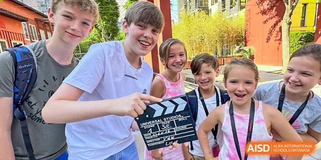 A group of young students smile while holding a film clapperboard during a creative media project. Deutsche Botschaftsschule Peking