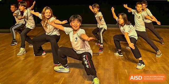 Four young students perform a synchronized martial arts or dance routine on a wooden stage at the Deutsche Botschaftsschule Peking.