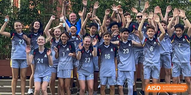 A large group of students in matching sports uniforms cheer and wave their hands at the Deutsche Botschaftsschule Peking.