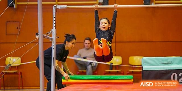 A young student performs a focused pike hang on the uneven bars while a coach assists with landing mats nearby. Deutsche Botschaftsschule Peking