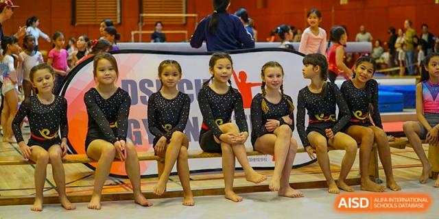 Five young gymnasts in matching black and gold leotards sit together on a bench during a competition break. Deutsche Botschaftsschule Peking