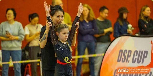 A coach helps a young gymnast maintain perfect form and posture during a training session in the sports hall. Deutsche Botschaftsschule Peking