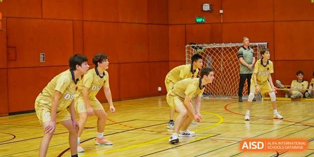 Three volleyball players in gold uniforms stand ready on the court, focusing intensely on the incoming serve. Deutsche Botschaftsschule Peking