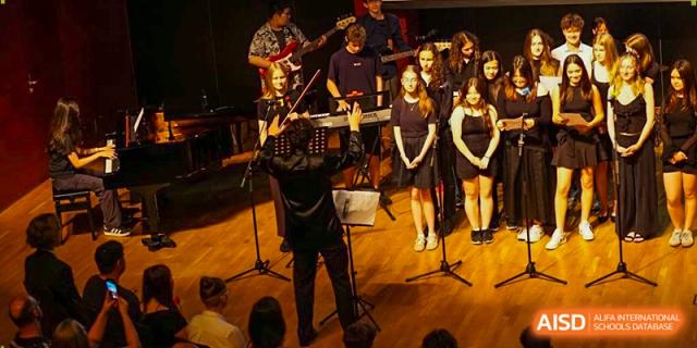 An orchestral conductor leads a group of student musicians and singers during an evening performance. Deutsche Botschaftsschule Peking