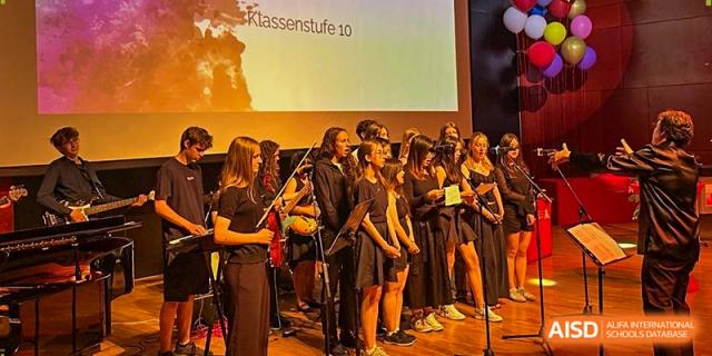 A student choir in black attire performs on stage under warm lighting during a musical showcase. Deutsche Botschaftsschule Peking