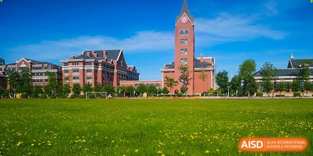 A tall red brick clock tower rises above a lush green lawn on a clear day at the center of Harbin Meijia Foreign Language School.