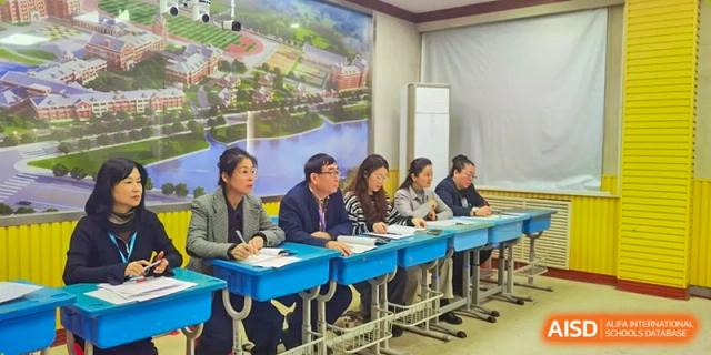 A panel of educators sits at a long blue table, taking notes and observing a demonstration in a bright classroom at Harbin Meijia Foreign Language School.