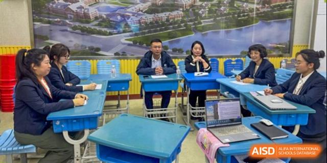Two staff members sit at school desks in front of a scenic wall mural during an indoor event at Harbin Meijia Foreign Language School.