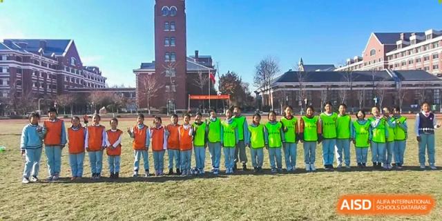 A group of young students in colorful green and orange athletic vests stand in a line on a grassy field in front of a large brick school building at Harbin Meijia Foreign Language School.