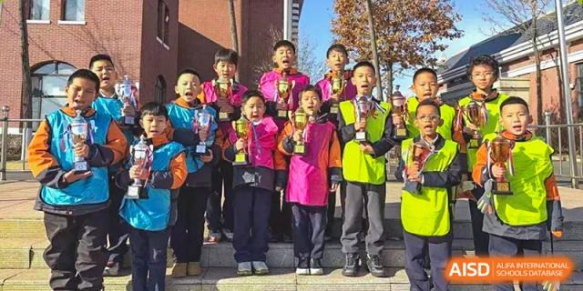 A group of joyful young students stands on outdoor steps, proudly holding up gold trophies after a successful sports competition at Harbin Meijia Foreign Language School.
