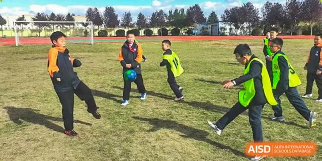 A young student in a black and orange jacket dribbles a blue soccer ball across a green field during practice at Harbin Meijia Foreign Language School.
