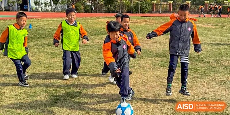 A young boy in an orange athletic jacket focuses on kicking a soccer ball during an outdoor physical education class at Harbin Meijia Foreign Language School.