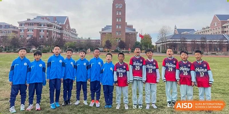 Students stand in a line on the sports field wearing various athletic jerseys with the brick school tower in the background at Harbin Meijia Foreign Language School.