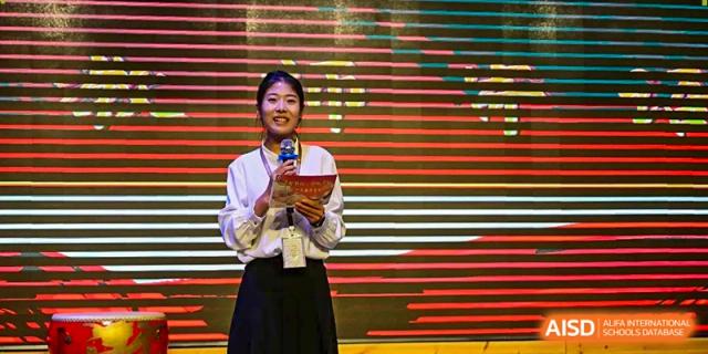 A young woman smiles while holding a program and speaking into a microphone on a stage with a vibrant digital backdrop at Harbin Meijia Foreign Language School.