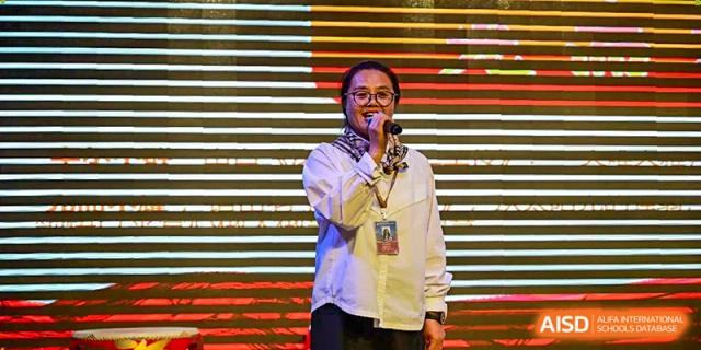 A female educator in a white shirt and patterned scarf speaks into a microphone during a school assembly at Harbin Meijia Foreign Language School.