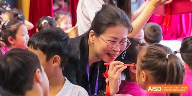 An instructor smiles warmly while assisting a student with a traditional craft during an indoor workshop at Harbin Meijia Foreign Language School.