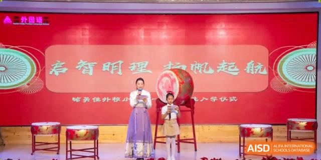 A student in traditional dress stands on a stage next to a large red drum during a formal school ceremony at Harbin Meijia Foreign Language School.