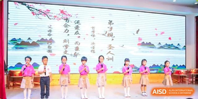 Four young girls in matching pink outfits stand in a row on stage and sing into microphones during a musical performance at Harbin Meijia Foreign Language School.