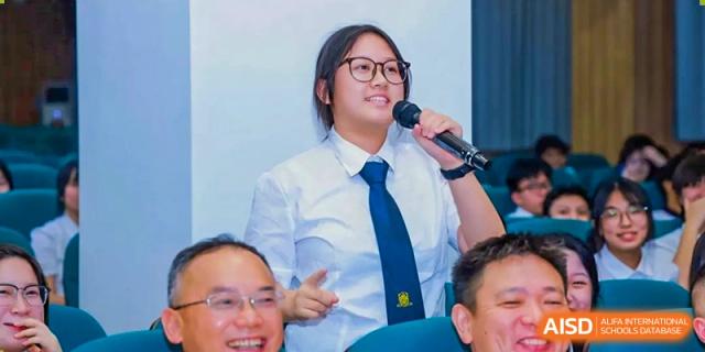 A female student in a white shirt and blue tie smiling while speaking into a microphone at Maple Leaf International Schools - Hainan