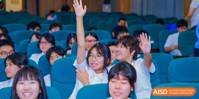 A female student raises her hand while sitting in a large audience during a presentation at Maple Leaf International Schools - Hainan