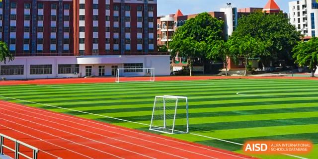 An outdoor view of a green soccer field and red running track next to a multi-story brick school building at Maple Leaf International Schools - Hainan