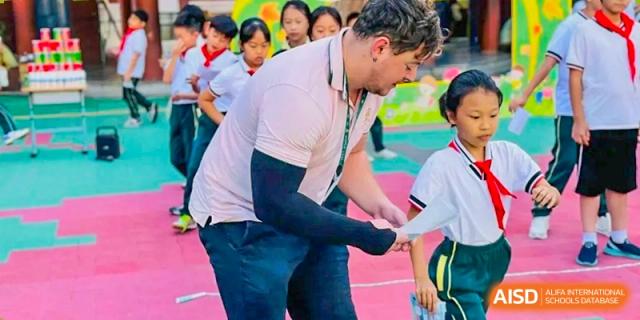 A male teacher in a light pink shirt leaning down to assist young students outdoors at Maple Leaf International Schools - Hainan