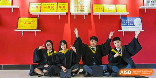 Three students in black graduation gowns posing with peace signs in front of a red wall with awards at Maple Leaf International Schools - Hainan