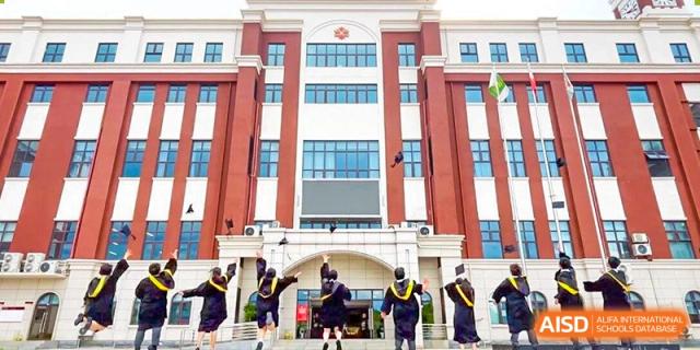 Four graduates in black gowns tossing their caps into the air in front of a red and white school building at Maple Leaf International Schools - Hainan