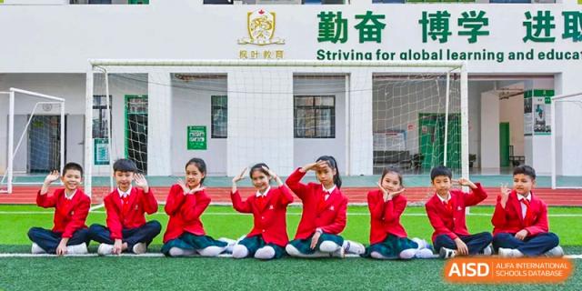 Four young students in red school blazers and plaid skirts pose playfully on an outdoor green sports field Maple Leaf International Schools - Hainan