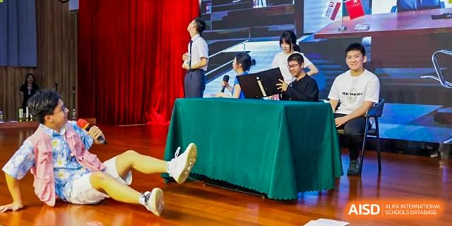 Students engage in a dramatic theater production on stage featuring props and a large green draped table Maple Leaf International Schools - Hainan