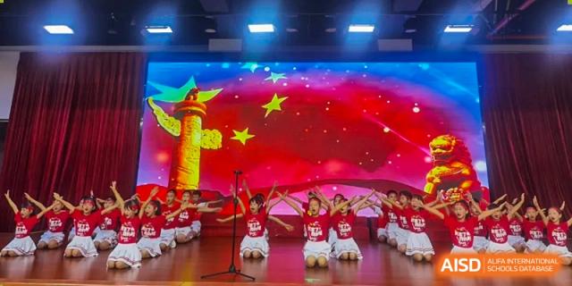 Children in red shirts kneeling and performing on a stage with a bright graphic background of yellow stars at Maple Leaf International Schools - Hainan
