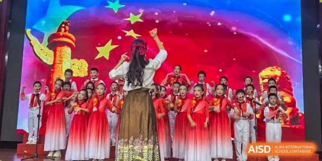 A conductor leads a choir of students dressed in red and white on a brightly lit stage at Maple Leaf International Schools - Hainan