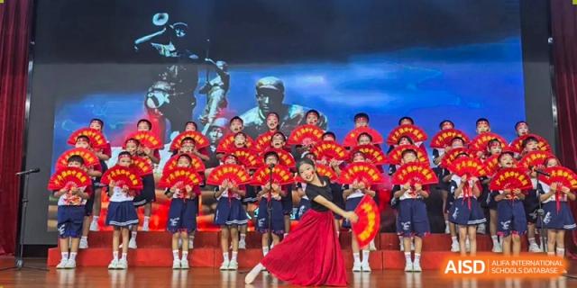 Students holding red fans and posing on stage in front of a blue backdrop featuring a historical mural at Maple Leaf International Schools - Hainan