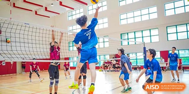 A student athlete in a blue jersey jumps high for a spike during a competitive volleyball match in the school gymnasium at Guangzhou ULink International School.
