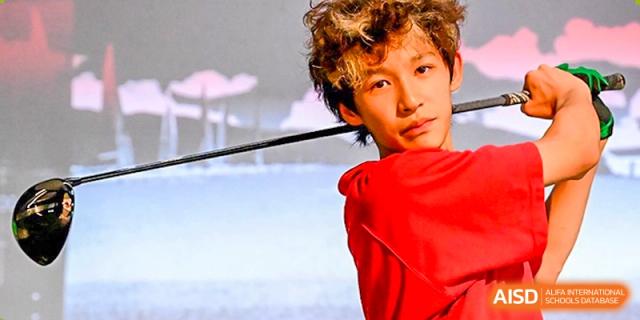A close-up portrait of a male student in a red uniform holding a golf club over his shoulder during an indoor practice session at Guangzhou ULink International School.