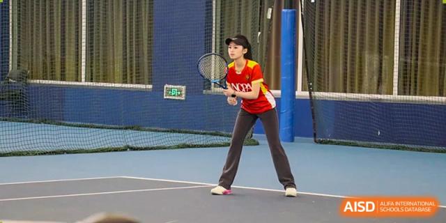 A female student in a red and yellow sports jersey prepares to return a shot on an outdoor tennis court at Guangzhou ULink International School.