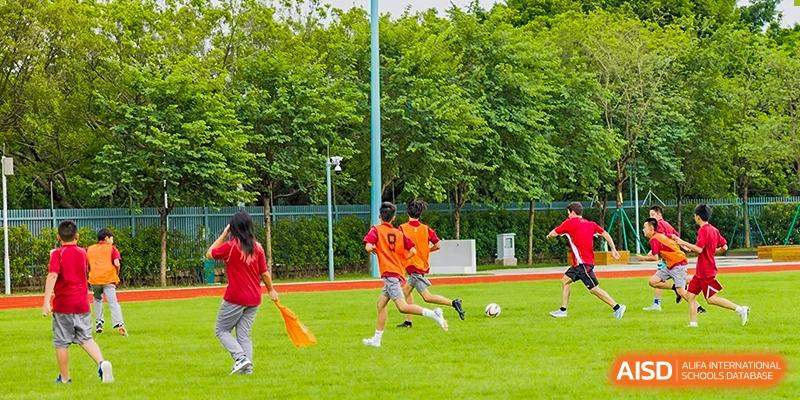 Students in orange jerseys participate in a soccer drill on a lush green field during an outdoor physical education class at Guangzhou ULink International School.