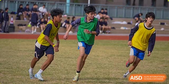 A male student wearing a green pinnie focuses on a soccer match on the grass field during an outdoor sports session at Guangzhou ULink International School.