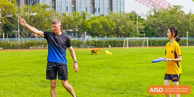 A sports coach or student standing on the edge of a lush green athletic field during a sunny outdoor training day at Guangzhou ULink International School.
